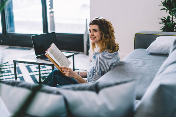 Cheerful young woman looking away while turning page of book sitting on floor.Positive hipster student holding bestseller in hands enjoying leisure time at home interior of modern apartment - Photo, image