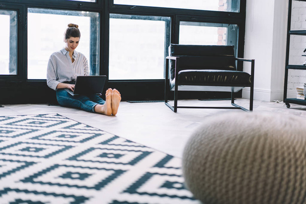Pensive young woman sitting on floor and working remotely at modern laptop using wireless internet connection in stylish apartment.Concentrated hipster girl searching information and installing app - Φωτογραφία, εικόνα