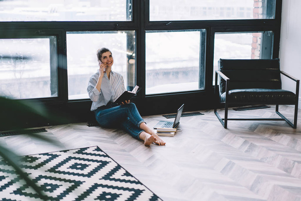 Positive young woman calling on digital smartphone device and talking with friend to plan meeting sitting on floor with notepad in modern apartment with stylish interior and big windows - Fotografie, Obrázek