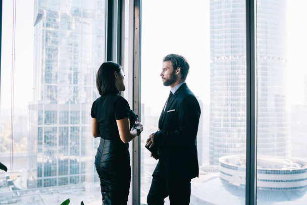 Male and female entrepreneurs dressed in elegant apparel communicating about projest ideas near window while waiting business partner for cooperation, confident proud ceo in office building - Foto, Imagem