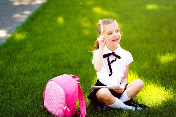 Bingo. Little school girl with pink backpack sitting on grass after lessons and thinking ideas, read book and study lessons, writing notes, education and learning concept - Фото, изображение