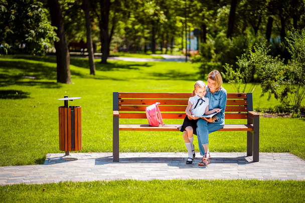 Mother and little daughter in outdoors public park sitting on the bench and read book, study lessons. Parenthood and child concept - Fotografie, Obrázek