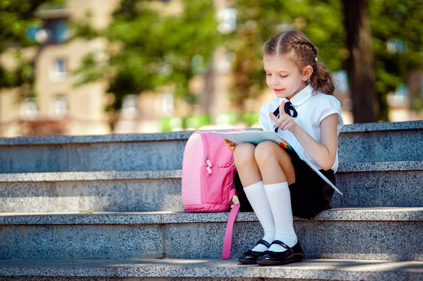 El alumno de la escuela primaria lee el libro sentado en las escaleras. Chica con mochila cerca del edificio al aire libre. Comienzo de las lecciones. Primer día de otoño
 - Foto, imagen