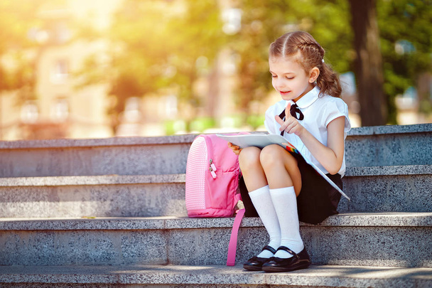 Pupil of primary school read book sitting on stairs. Girl with backpack near building outdoors. Beginning of lessons. First day of fall - Foto, afbeelding