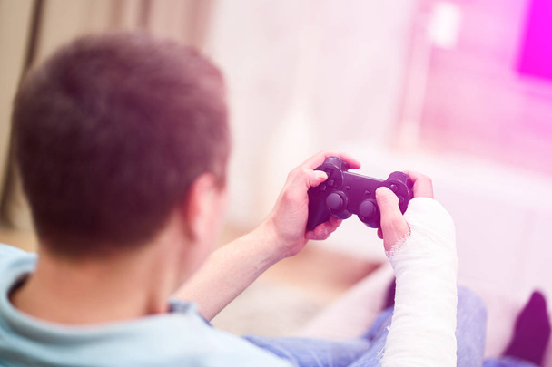 Man with broken arm in plaster cast holding controller and playing in videogame in front of TV. Game addiction concept. - Foto, Imagen