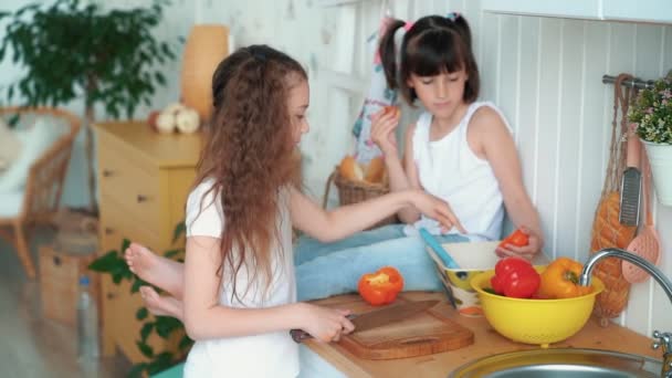 Girl cuts pepper in kitchen, her sister sits on table and eats bun, slow motion - Metraje, vídeo