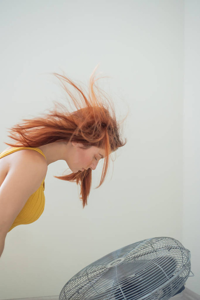 Portrait of a beautiful red-haired woman in mustard overalls enjoying the cooling breeze from a large electric fan. The girl is refreshed in the summer sweltering heat. Hair develops from the wind. - Foto, afbeelding