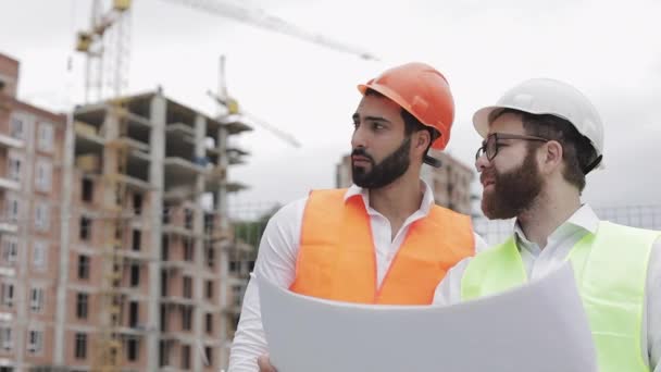 Male construction engineer discussion with architect at construction site or building site of highrise building. They holding construction drawings in their hands. - Video