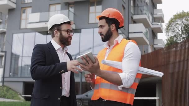 Construction manager and architect man are discussing the construction plan of the modern business center. They checks the work with a tablet. Outside. - Filmati, video
