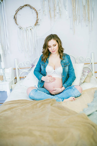 Piękna kobieta w ciąży w niebieskie dżinsy jest na łóżku. Koncepcja szczęśliwego macierzyństwa, zdrowego stylu życia - Zdjęcie, obraz