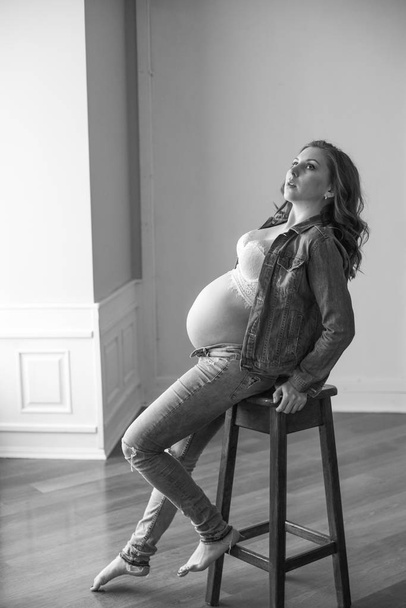 Beautiful pregnant woman in blue jeans and denim jacket  on a high wooden chair. Concept of happy motherhood, healthy lifestyle - Fotografie, Obrázek