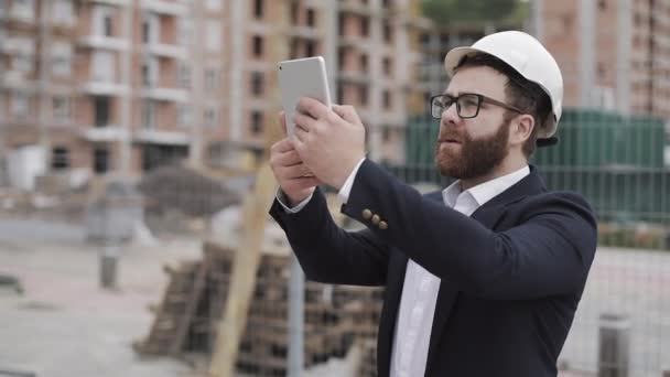 Young bearded architect at the construction site taking photo with tablet. He dressed in business suit. - Footage, Video