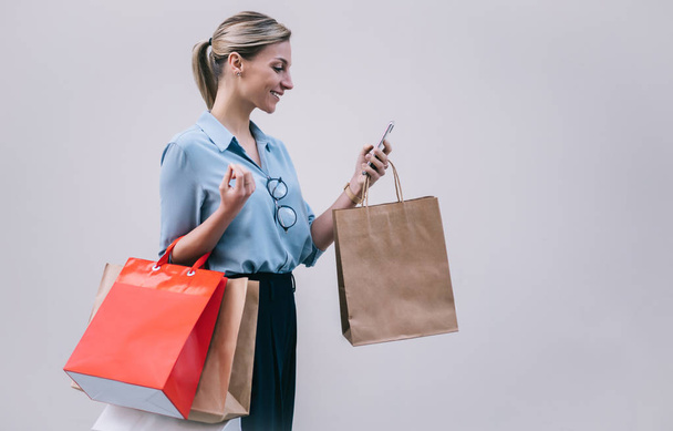 Cheerful smiling girl carrying paper bags standing near promotional background after shopping feeling good from received text message with sale discounts information, happy woman browsing internet - Foto, Bild