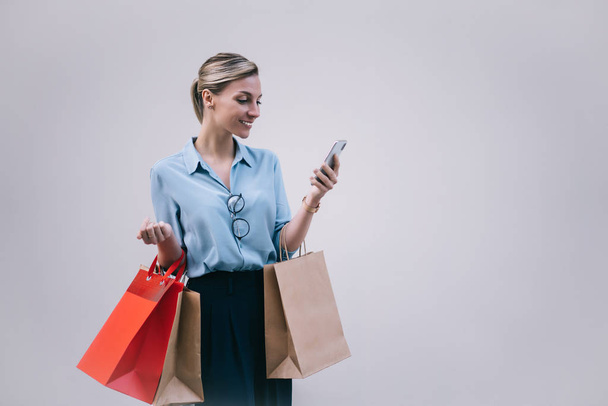 Positive caucasian shopper in stylish wear holding paper bags and enjoying online messaging via smartphone application, happy hipster girl chatting near promotional background on publicity area - Foto, afbeelding