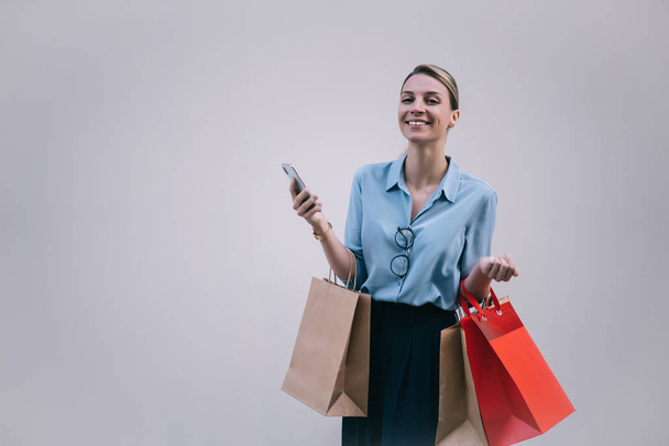 Half length portrait of cheerful female shopper with paper bags and mobile phone in hands feeling excited from Black Friday sales, happy hipster girl smiling at camera near copy space area for text - Foto, Bild