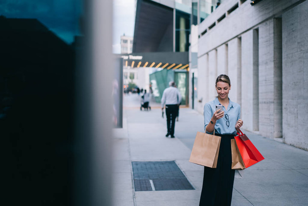 Happy kobieta shopper z papierowymi torbami w ręku korzystających rekreacji po zakupie zakupów dokonywania rezerwacji online dla taksówki, wesoły hipster dziewczyna czatując z przyjacielem smsowania wiadomości o sprzedaży - Zdjęcie, obraz