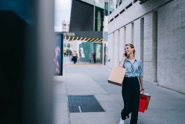 Stylish young woman calling on smartphone while strolling in mall with paper bags after good shopping, hipster girl bought casual clothes in stores and talking on mobile phone about Black Friday - Foto, Imagen