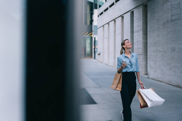 Cheerful woman with paper bags and smartphone in hands strolling on street and looking for branded shop with big sales for customers, stylish female buyer with packages enjoying leisure time - Фото, изображение