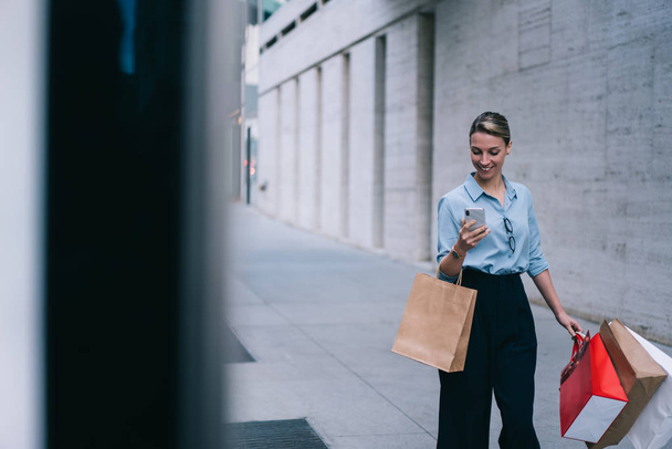 Cheerful female fashion blogger checking messages and comments from followers at own website connected to public internet while walking in downtown and searching brand store with sales on Black Friday - Фото, зображення