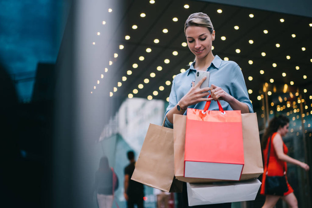 Happy female customer holding paper bags after buying purchase in mall during Black Friday, positive caucasian woman shopaholic resting after shopping time while waiting friend on publicity area - Photo, image