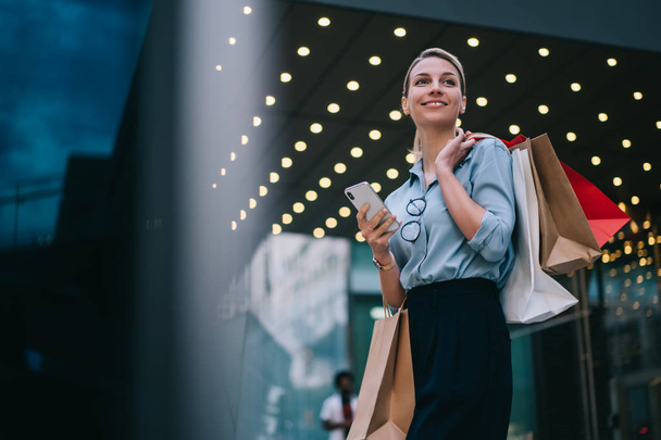 Good looking caucasian customer holding mobile standing near mall doing shopping in black friday, cheerful caucasian customer with paper bags using public internet connection on smartphone gadget - 写真・画像