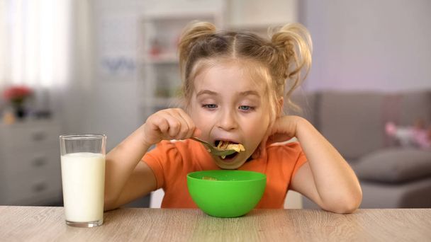 Happy little girl eating cornflakes with milk sitting home table, healthy food - Photo, Image
