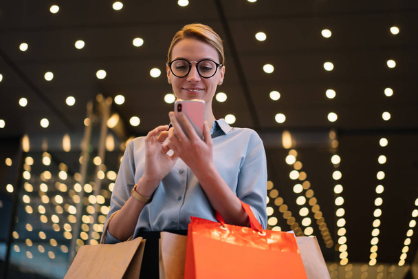 Smiling female customer in eyewear standing outdoors with shopping bags and using wireless 4g internet on smartphone device for installing taxi application, concept of technology and communication - Zdjęcie, obraz