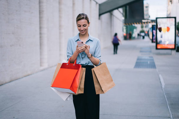 Cheerful caucasian woman blogger posting publication in social network using 4g internet connection on mobile phone, happy hipster girl with shopping packages messaging with friend via smartphone - Zdjęcie, obraz