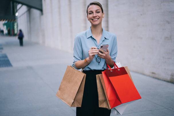 Half length portrait of excited female buyer standing on publicity area with paper packages and smiling at camera, happy hipster girl in trendy apparel holding modern cellular gadget outdoors - Valokuva, kuva