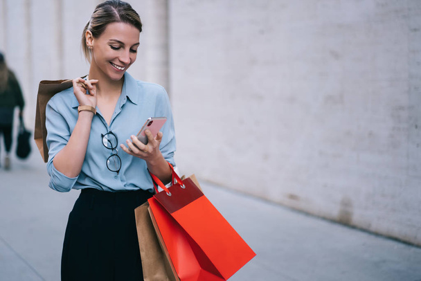 Девушка-хипстер читает сообщение в социальной сети через смартфон, стоя на городской местности, улыбается покупательнице и ведет блог с помощью мобильного телефона после посещения магазинов с распродажами и скидками - Фото, изображение
