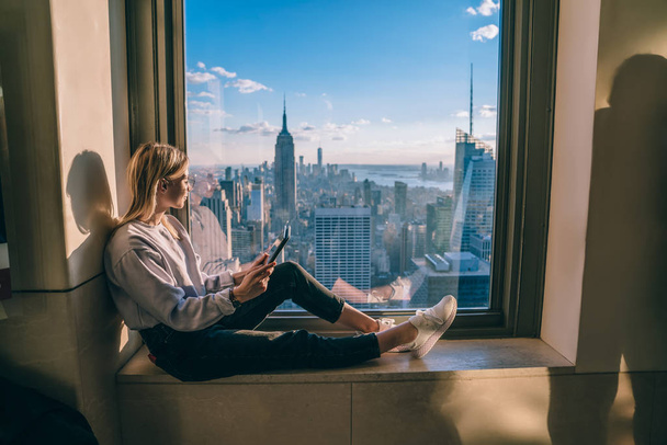 Caucasian hipster girl using modern tablet while sitting on window sill with breathtaking metropolis cityscape view on New York. Female traveler enjoying scenery Empire State Building from coworking - Фото, зображення