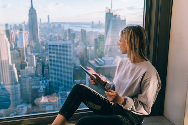 Stylish blonde female in casual wear observing scenic views of Empire State building landmark and cityscape downtown while sitting in window sill. Popular attractions of New York City. Digital nomad - Foto, imagen