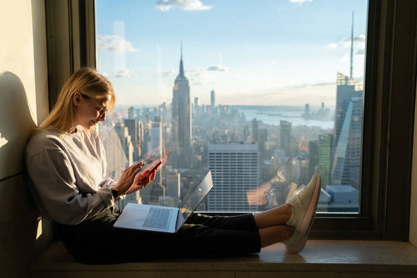 Trendy caucasian female texting message on smartphone while sitting on office window sill with laptop computer. Young student hipster blogger using application on mobile phone.Exchange abroad New York - Φωτογραφία, εικόνα