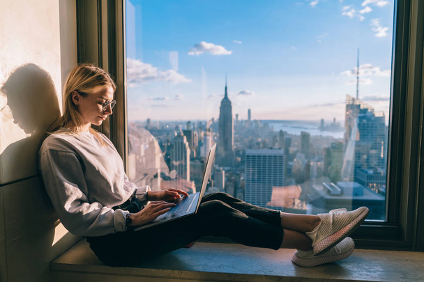 Concentrated female freelancer keyboarding information on laptop computer working remote in coworking office with panoramic views of scenic Manhattan downtown. Student exchange and travel to New York - 写真・画像