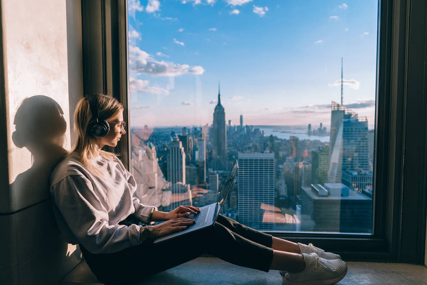 Caucasian female listening to music in headphones while working on laptop computer and enjoying inspiring beautiful view of metropolitan New York city from skyscrapers window. Hipster girl tourist - Zdjęcie, obraz