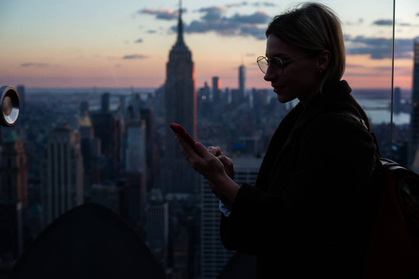 Hipster girl in autumn coat using smartphone roaming network while standing on Observation deck with scenery Empire State building view.Female tourist publishing fresh photos for travel lifestyle blog - Foto, afbeelding