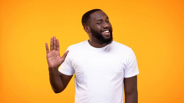 Affable young black man showing palm, waving hand neighborly yellow background - Fotó, kép