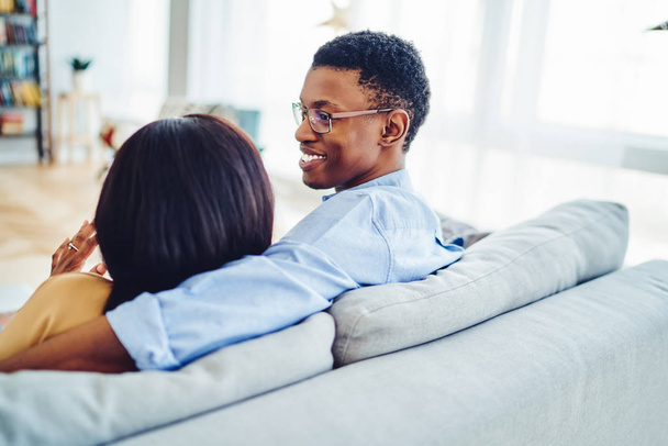 Smiling african american male looking at his girlfriend with love having talk while relaxing in living room, rear view of dark skinned couple recreating at weekends at home interior sitting on sof - Photo, image