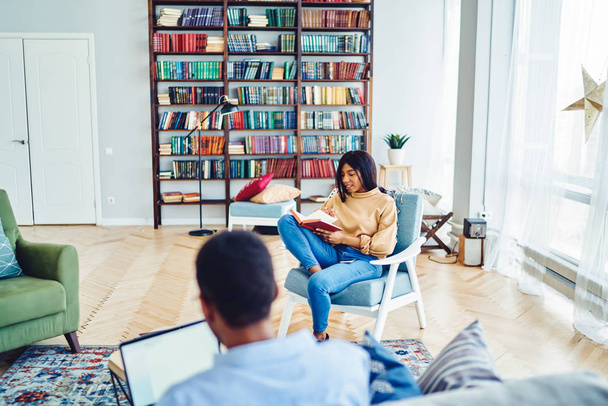 Young dark skinned hipster students spending time in hostel during weekends recreation, african american woman sitting in chair reading interesting literature from home book collection in living roo - 写真・画像