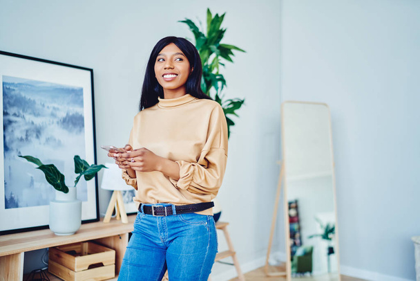 Cheerful african american female using mobile phone for chatting spending time at home interior with wifi, smiling dark skinned hipster girl holding smartphone making booking spending time at fla - Foto, Bild