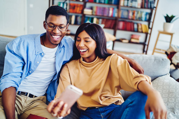 Cheerful dark skinned hipster couple making funny faces posing for selfie and using smartphone app, happy african american girl and guy sitting on sofa spending time at home making picture togethe - 写真・画像