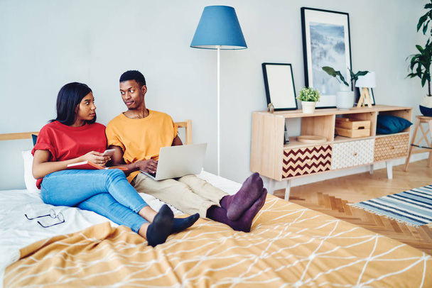 African american couple in love talking to each other sitting at bed room using laptop computer and smartphone for networking, dark skinned young family having conversation resting on free tim - Fotó, kép