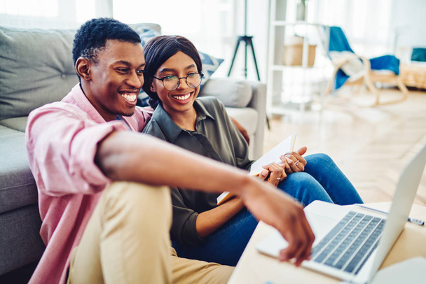 Cheerful african american couple satisfied with resting together in cozy interior living room watching movie on laptop computer, smiling dark skinned male and female laughing at funny video - Foto, imagen