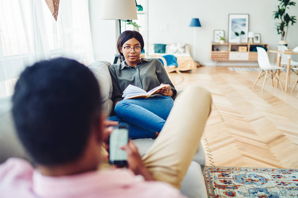 Πορτρέτο της αφροαμερικάνας γυναίκας με γυαλιά που κάθεται με το βιβλίο της να αναπαριστά τον καναπέ με το αγόρι της, μελαμψό γάμο, περνώντας ελεύθερο χρόνο στο διαμέρισμα στις διακοπές και τη λεϊσούρ - Φωτογραφία, εικόνα