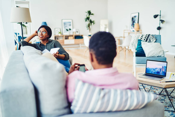 Dreamy smiling african american girl looking away holding book recreating on sofa at apartment with boyfriend, dark skinned couple spending free time at modern designed living room with stylish deco - Φωτογραφία, εικόνα