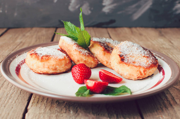 Gourmet breakfast - cottage cheese pancakes, cheesecakes, cottage cheese pancakes with strawberries, mint and powdered sugar in a white plate. Useful dessert on a wooden table in rustic style. Selective focus. - Photo, image
