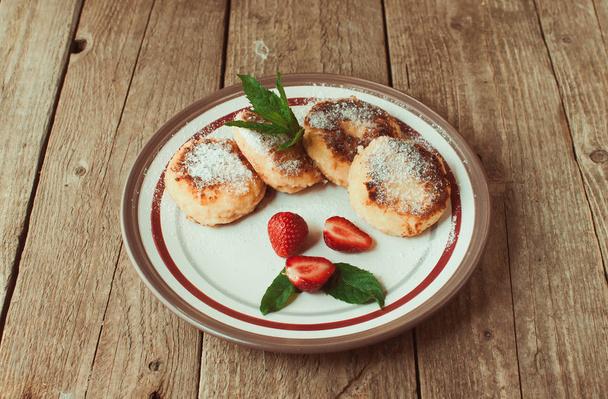 Gourmet breakfast - cottage cheese pancakes, cheesecakes, cottage cheese pancakes with strawberries, mint and powdered sugar in a white plate. Useful dessert on a wooden table in rustic style. Selective focus. - Foto, imagen