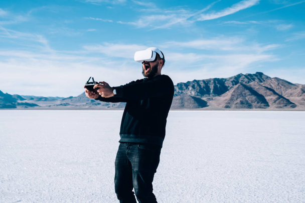 Amazed male controlling with joystick virtual reality dimension while wearing VR headset outdoors in empty nature cosmic landscape. Excited man playing interactive futuristic game app with gamepad  - Foto, Imagen