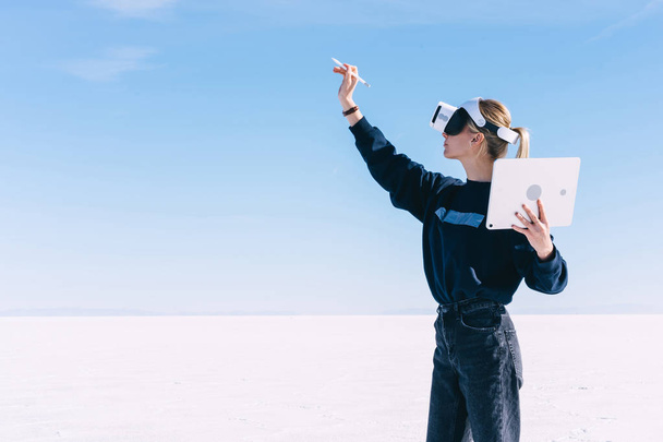 Young women extends hand with digital pencil for interactive touch over air while standing in spacious clear outdoors space wearing virtual reality headset. Hipster girl designing architectural space - 写真・画像