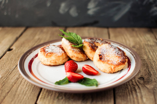 Gourmet breakfast - cottage cheese pancakes, cheesecakes, cottage cheese pancakes with strawberries, mint and powdered sugar in a white plate. Useful dessert on a wooden table in rustic style. Selective focus. - Фото, зображення